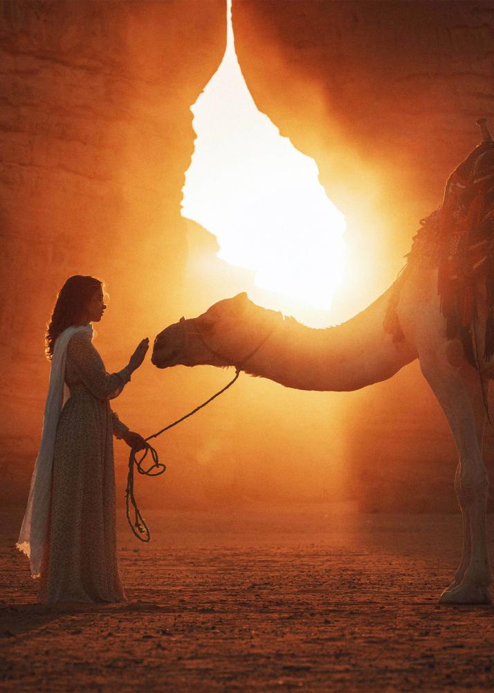 Eine Frau in einem langen Kleid, die ein Kamel in einer Wüstenlandschaft bei Sonnenuntergang streichelt.