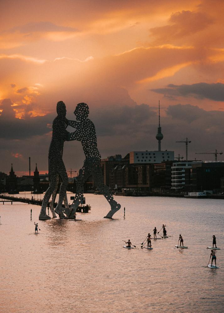 Eine große Skulptur steht im Wasser, während Menschen im Vordergrund auf Paddelbrettern fahren.