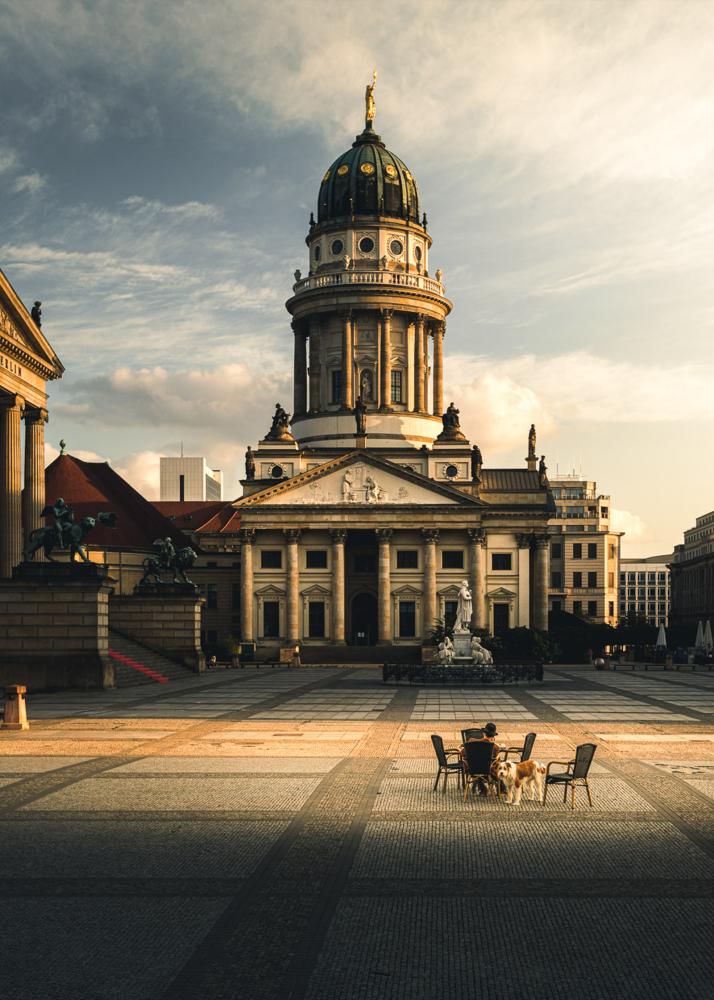 Zwei Personen sitzen mit einem Hund vor einem historischen Gebäude mit einer Kuppel.