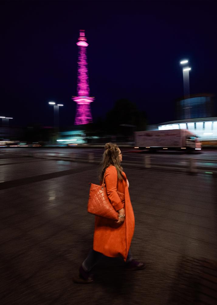 Eine Frau in einem orangen Mantel geht am pink beleuchteten Funkturm vorbei.