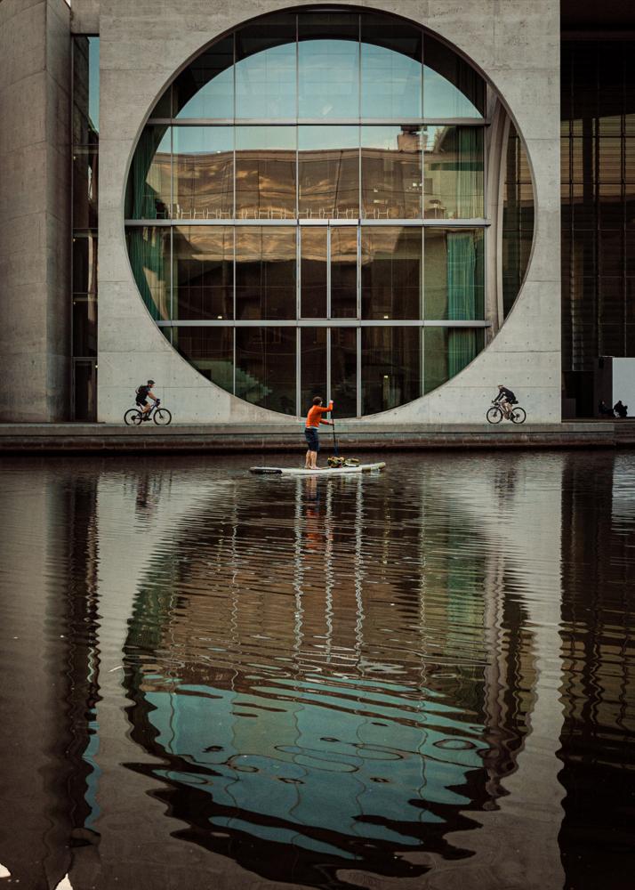Ein Mann steht auf einem Paddelbrett vor einem großen Gebäude mit runder Fensterfront und Wasserreflexionen.
