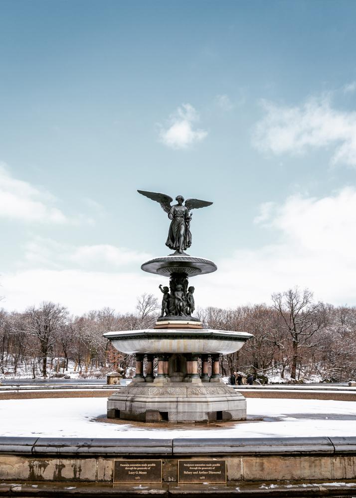 Ein Brunnen mit einer Engelstatue in der Mitte, umgeben von schneebedeckten Bäumen und einem klaren Himmel.