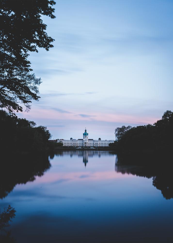 Ein Schloss am Wasser, das sich im ruhigen See spiegelt, umgeben von Bäumen bei Sonnenuntergang.