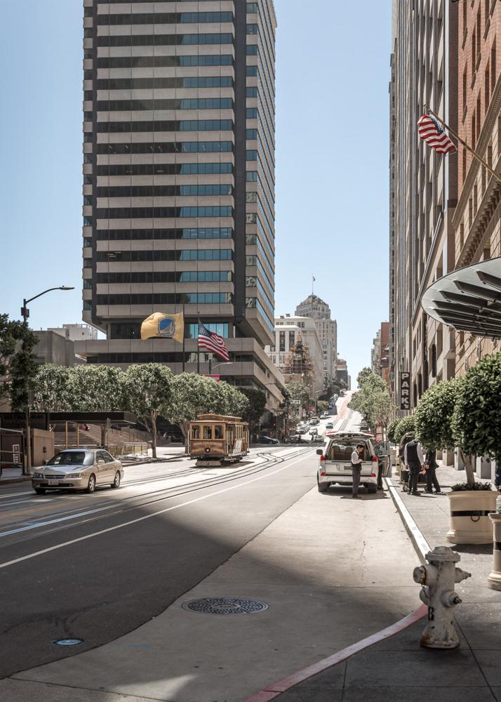 Eine Stadtstraße mit einer Straßenbahn, die zwischen hohen Gebäuden und Bäumen entlangfährt.