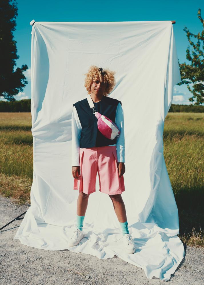 Eine Person mit lockigem Haar, in einem bunten Outfit mit pinkfarbenen Shorts und einer weißen Bluse, steht vor einem weißen Tuch in einer ländlichen Umgebung.