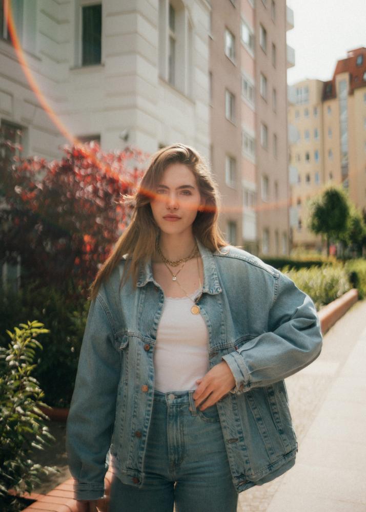 Eine junge Frau in einer Denim-Jacke und Jeans steht auf einem Gehweg mit einem Gebäude und Büschen im Hintergrund.