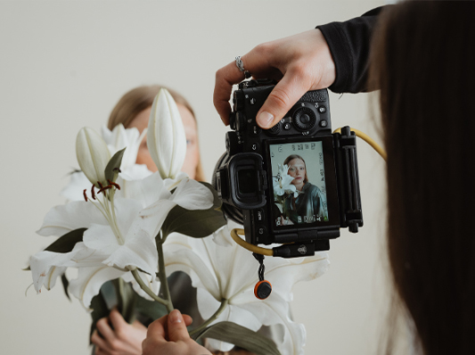 Auf diesem Bild sieht man im Vordergrund eine Sony Kamera und Blumen. Im Hintergrund ist ein Model welches gerade fotografiert wird.