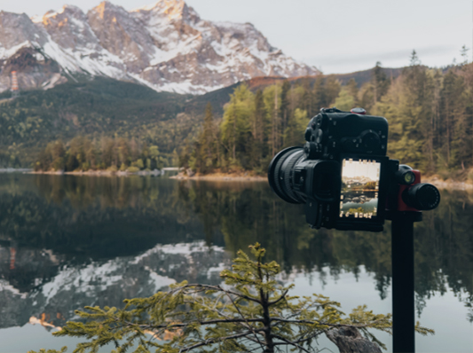 Auf diesem Bild sieht man im Vordergrund eine Sony Kamera auf einem Stativ. Im Hintergrund ist eine schöne Naturkulisse mit einem See und Bergen zu sehen.