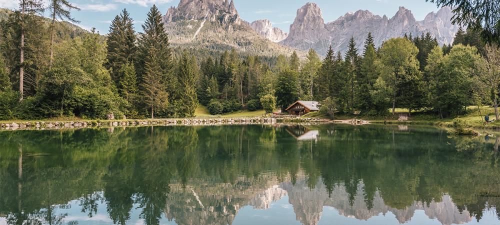 Ein ruhiger Bergsee spiegelt die umliegenden Bäume und schneebedeckten Berge perfekt wider, mit einem kleinen Haus am Ufer.