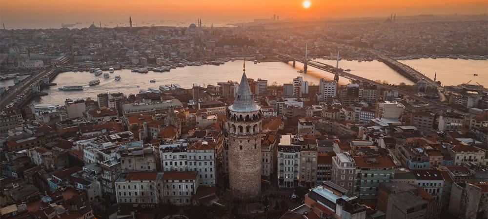 Eine Stadtlandschaft bei Sonnenuntergang mit einem markanten Turm im Vordergrund und einer Brücke, die über ein Gewässer führt.