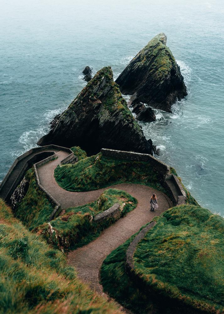 Ein steiniger Pfad führt zu felsigen Klippen am Meer, mit einer Person, die auf dem Weg steht und die Aussicht genießt.