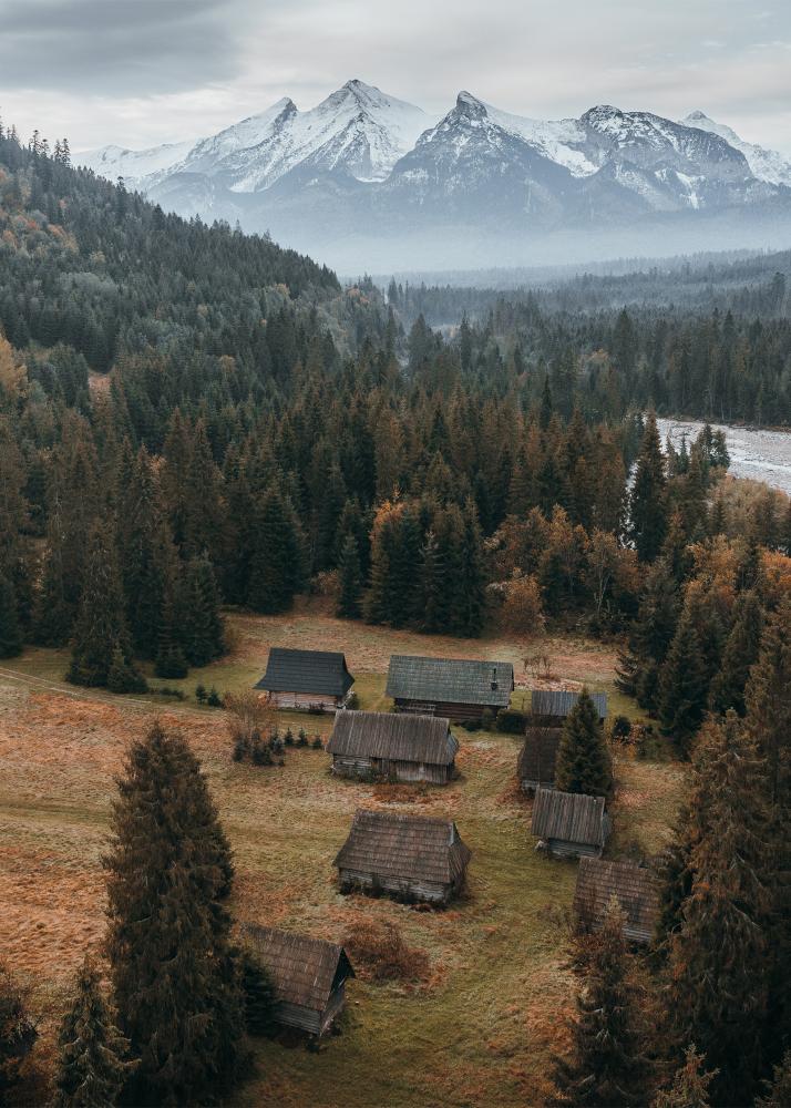 Eine ländliche Szene mit alten Holzhütten inmitten eines Waldes, mit schneebedeckten Bergen im Hintergrund.