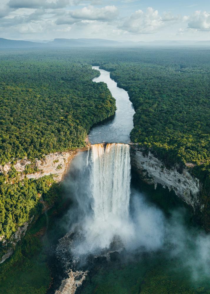 Ein majestätischer Wasserfall stürzt von einer Klippe in einen Fluss, umgeben von dichtem, grünem Wald und weitläufigen Bergen im Hintergrund.