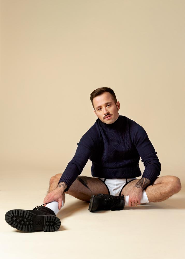 Ein Mann sitzt in entspannter Pose auf dem Boden vor einem beigen Hintergrund.