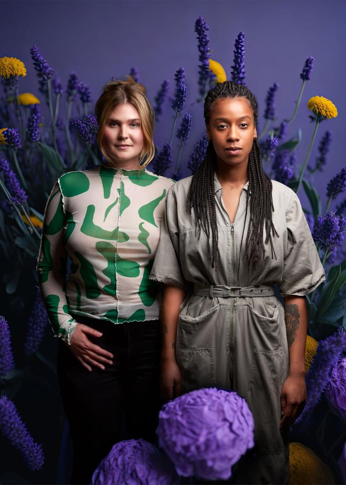 Zwei Personen stehen neutral und gerade, während sie direkt in die Kamera schauen. Sie sind umgeben von hohen, bunten Blumen.