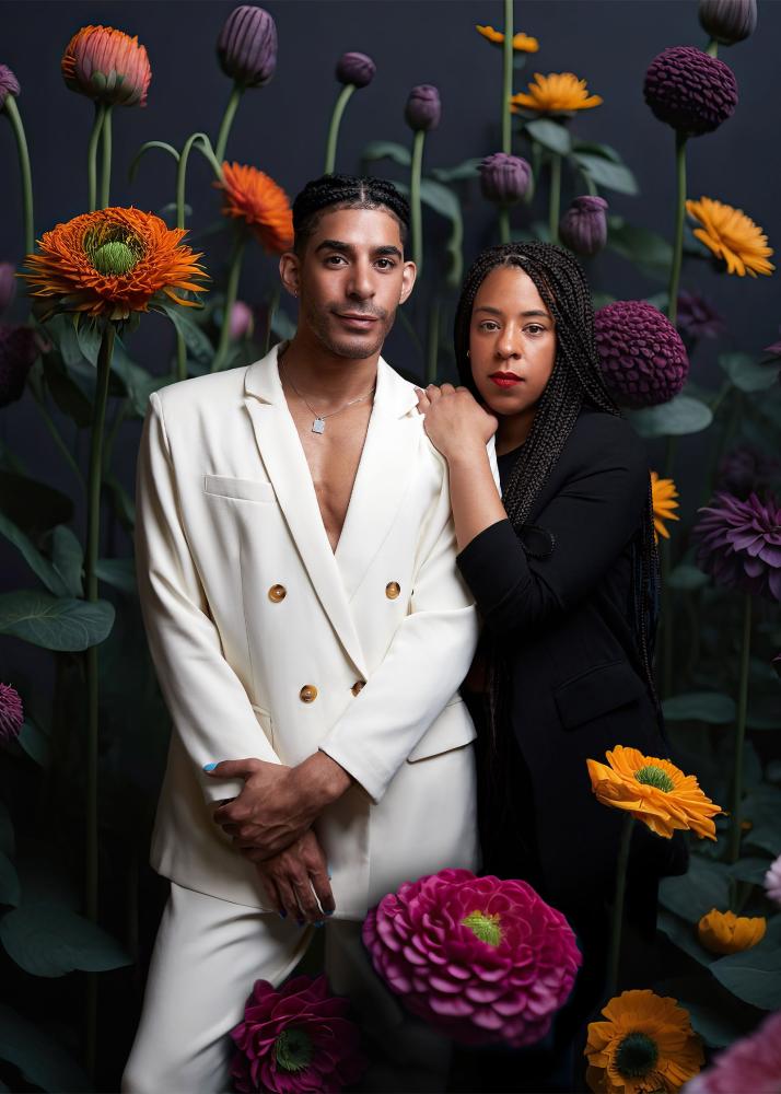 Zwei gut gekleidete Personen stehen in einem Feld mit hohen, bunten Blumen und schauen direkt in die Kamera.