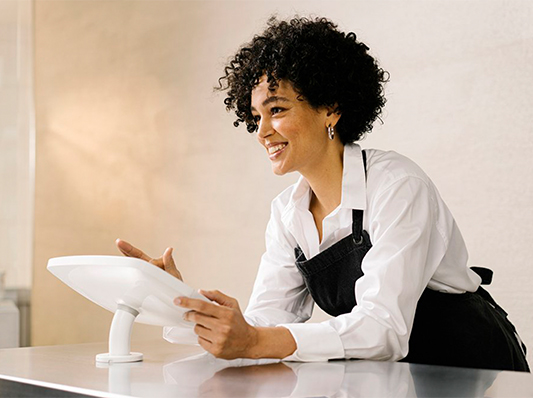 Eine Frau mit kurzem, lockigem Haar und einem schwarzen Schürzenkleid bedient ein Tablet in einem hellen Raum.