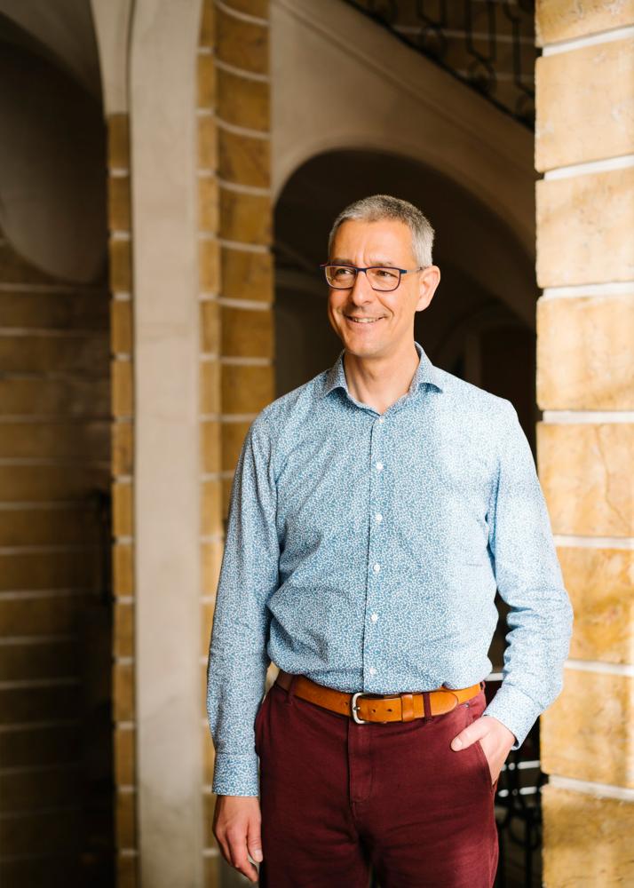 Ein Mann steht lächelnd in einem Gewölbegang, trägt ein blaues Hemd und eine braune Hose.