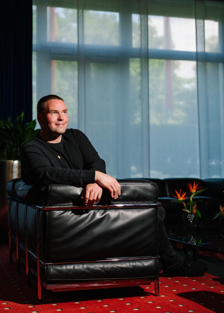 Ein Mann sitzt lässig in einem schwarzen Ledersessel in einem Raum mit großen Fenstern, durch die natürliches Licht einfällt.