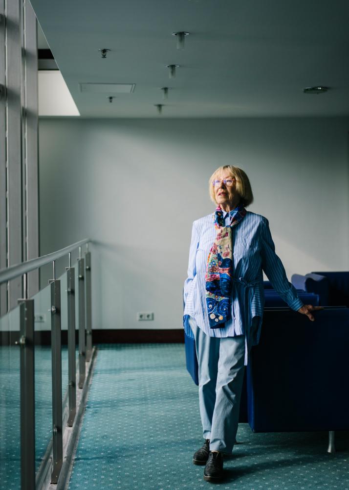 Eine ältere Frau geht durch einen hellen, modernen Innenraum mit großen Fenstern und einem Handlauf an der Wand.