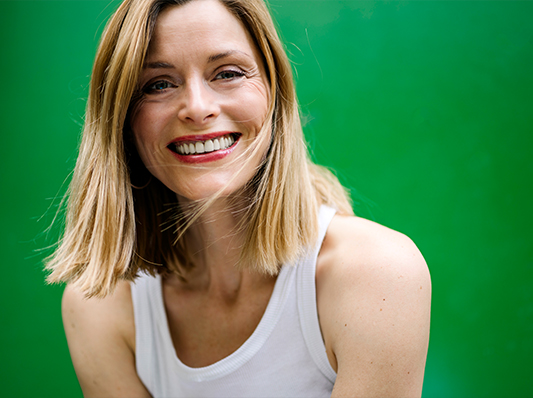 Eine Frau mit blondem Haar lächelt freundlich in die Kamera, vor einem grünen Hintergrund.
