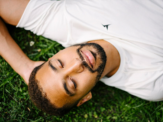 Ein Mann liegt entspannt auf dem Rücken im Gras und genießt die Sonne mit geschlossenen Augen.