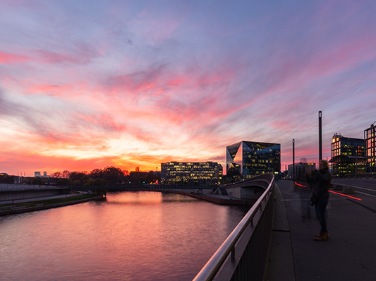 Ein Fluss bei Sonnenuntergang mit einer Brücke und modernen Gebäuden, die sich im Wasser spiegeln.