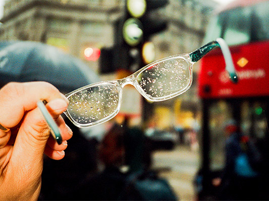 Eine Hand hält eine Brille mit Regentropfen vor einem roten Bus.