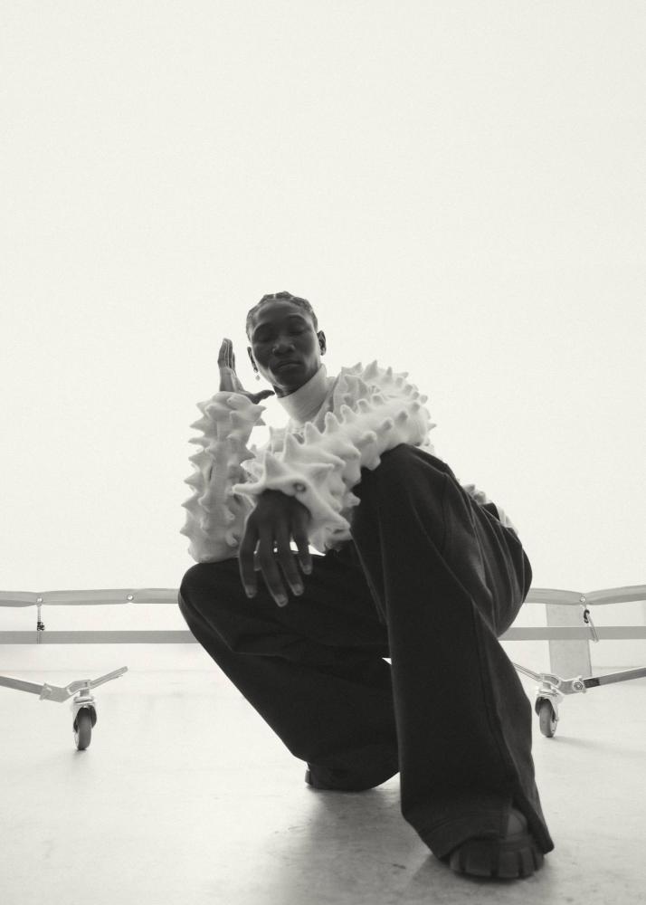 Ein Mann in einem strukturierten, weißen Pullover kniet auf einem Stuhl mit einer Hand an der Wange.