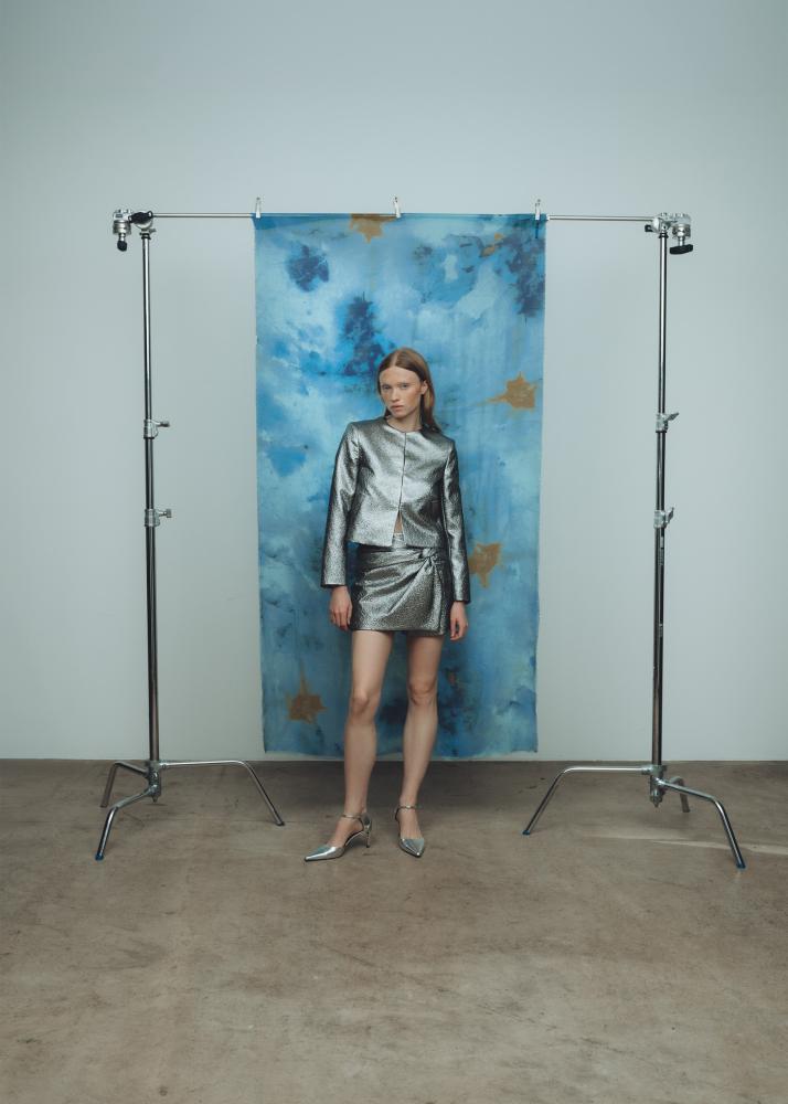 Eine Frau in einem glänzenden, silbernen Outfit steht vor einem blauen Hintergrund mit abstrakten Mustern.