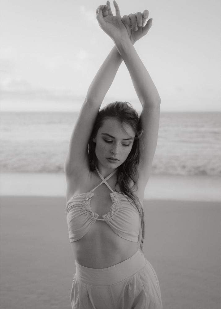 Eine Frau in einem weißen Bikini-Oberteil steht am Strand und streckt die Arme aus.