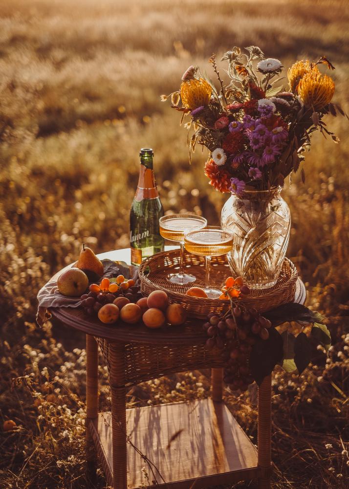 Ein Tisch im Freien mit einer Flasche, zwei Gläsern, einer Vase mit Blumen und einer Schale mit Früchten, umgeben von einem Feld.