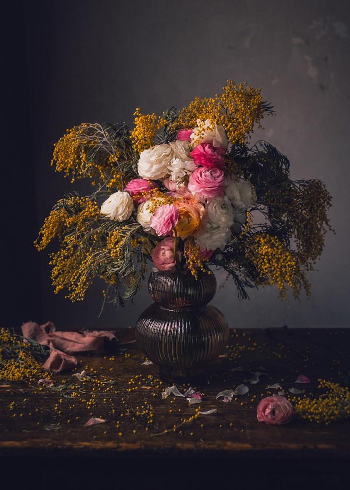 Ein Brautstrauß mit weißen, rosa und gelben Blumen in einer dunklen Vase auf einem Holztisch.