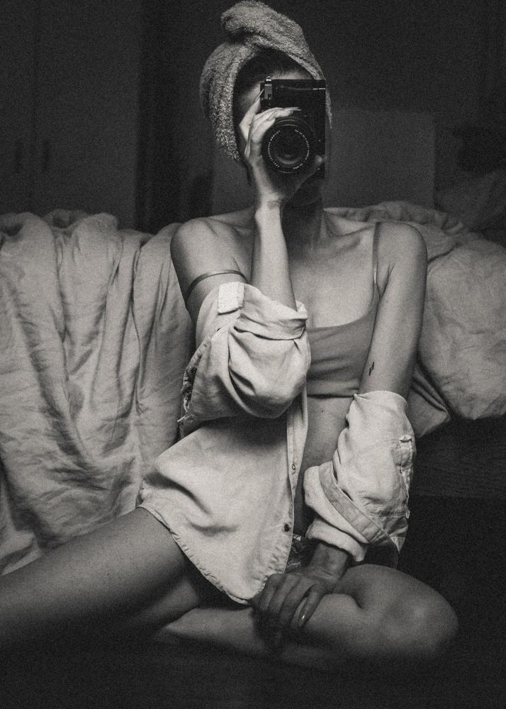 Eine Frau mit einem Handtuch-Turban hält eine Kamera vor ihr Gesicht, während sie vor einem Bett sitzt.