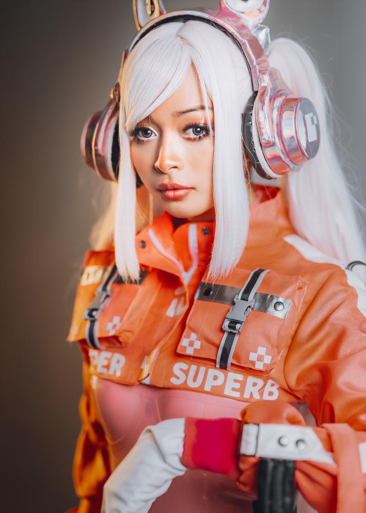 Eine Frau mit weißem Haar und futuristischem Kopfhörer in einem orangefarbenen Kostüm, das mit dem Wort 'SUPERB' bedruckt ist.