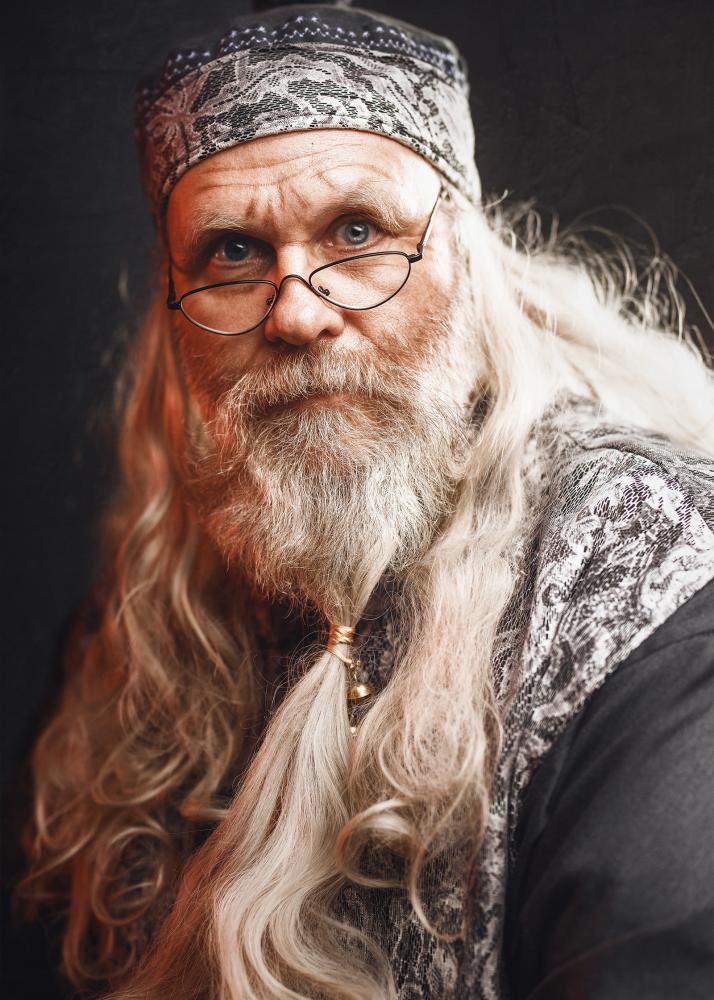 Ein Mann mit langem, weißem Haar und Bart, der eine Brille trägt und in ein verziertes Gewand gekleidet ist.