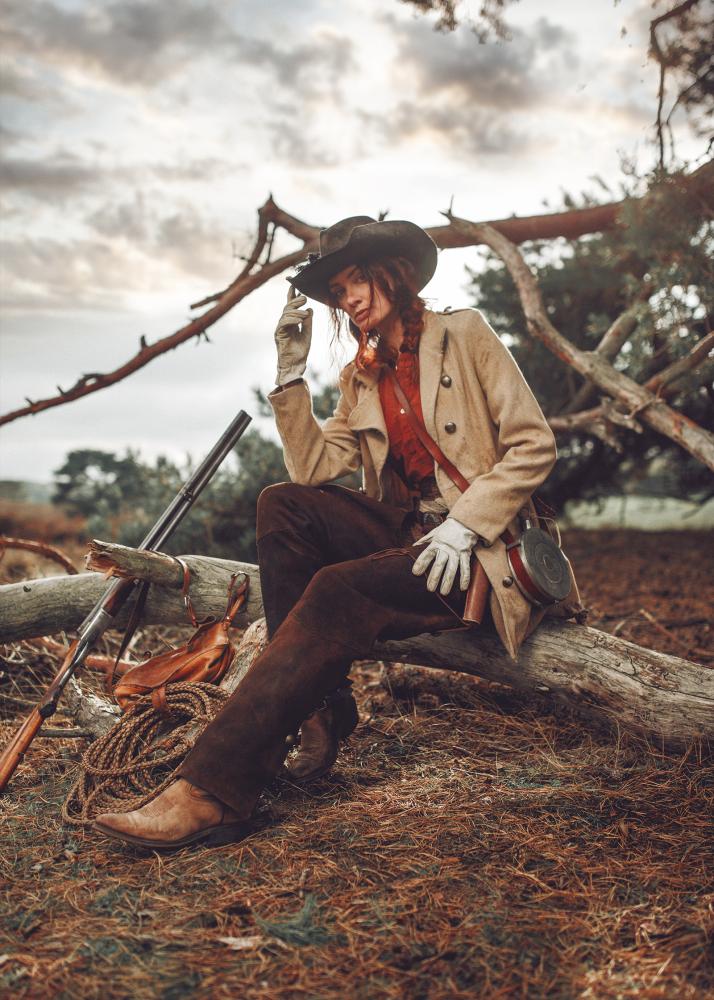 Eine Frau in einem Western-Outfit sitzt auf einem Baumstamm mit einem Gewehr und einem Hut.
