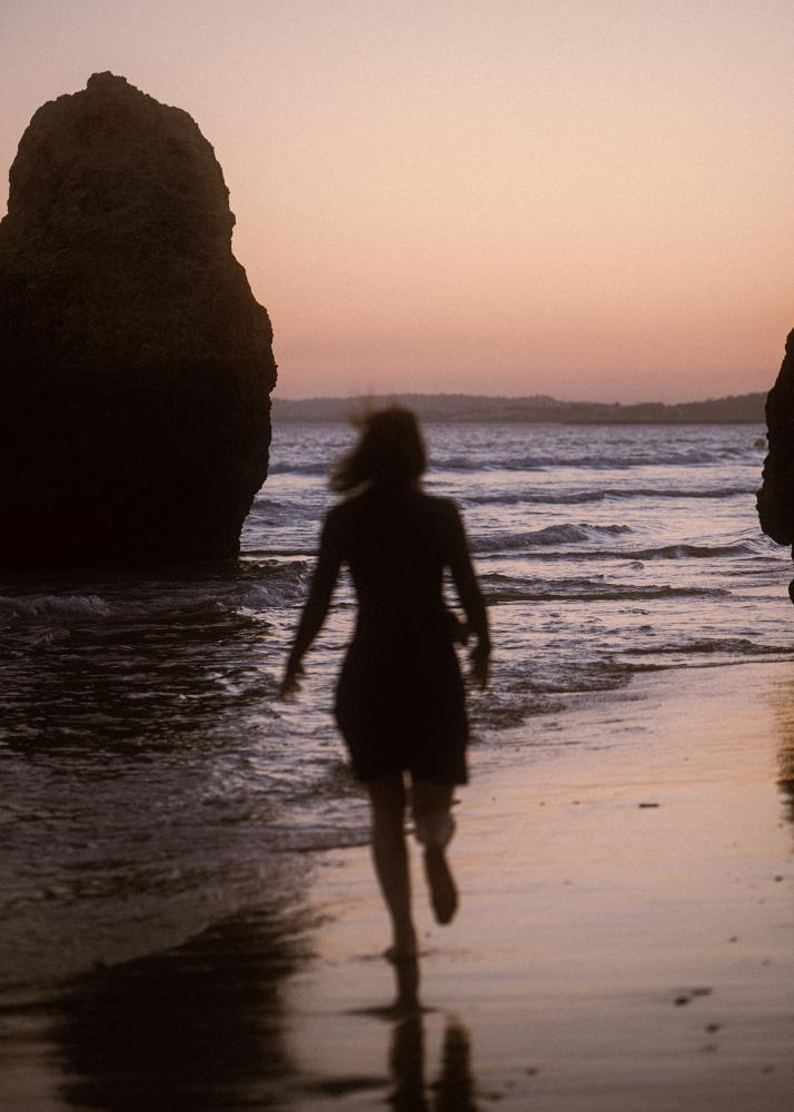 Eine Silhouette einer Frau, die am Strand entlangläuft, mit Felsen im Hintergrund und dem Sonnenuntergang.