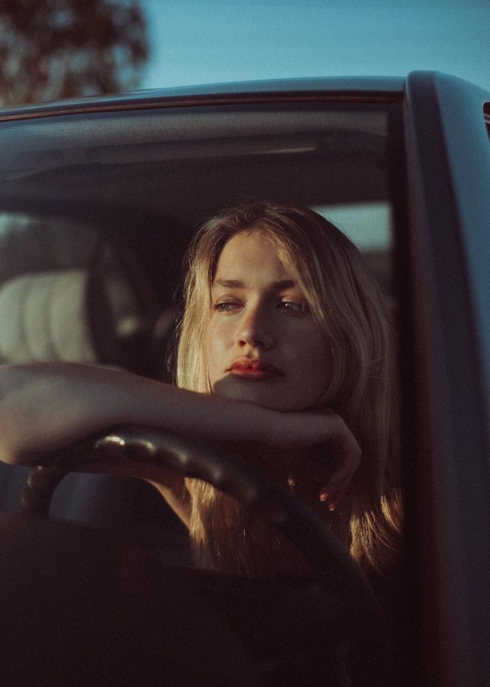 Eine Frau mit blondem Haar sitzt im Fahrerbereich eines Autos und hält das Lenkrad, während sie aus dem Fenster schaut.