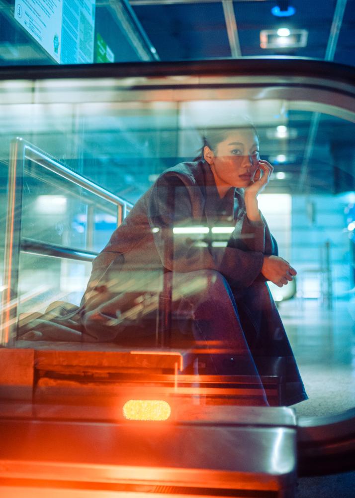 Eine Frau sitzt in einem öffentlichen Bahnhof und stützt ihren Kopf auf ihrer Hand ab, während sie aus durch die Glas-Balustrade einer Rolltreppe in die Kamera schaut.