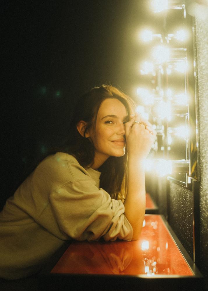 Eine Frau lehnt sich an einen beleuchteten Spiegel in einem Raum mit grellen Lichtern.