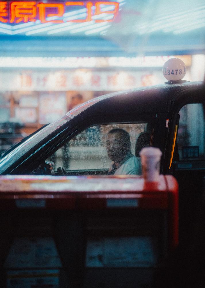 Ein Taxifahrer fährt durch eine beleuchtete Stadtstraße mit Neonlichtern im Hintergrund.
