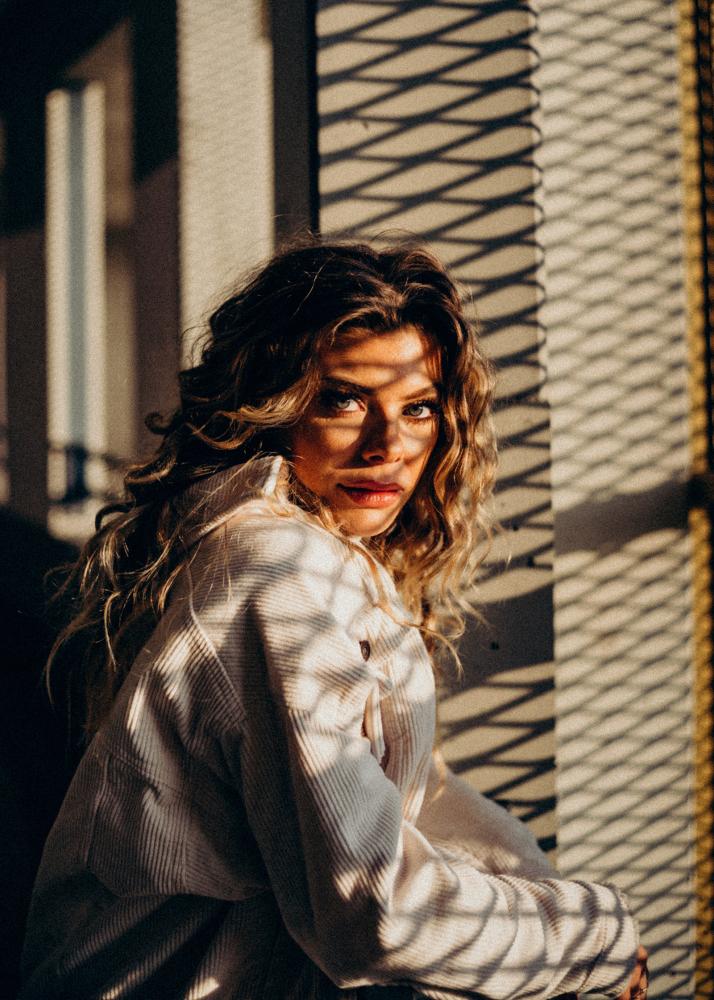 Eine Frau mit lockigem Haar lehnt an einem Metallzaun, während die Sonne durch die Gitter scheint.