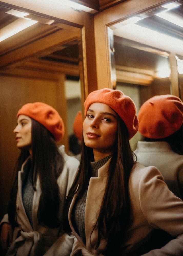 Eine Frau mit einem orangefarbenen Barett steht vor einem Spiegel in einem beleuchteten Raum.