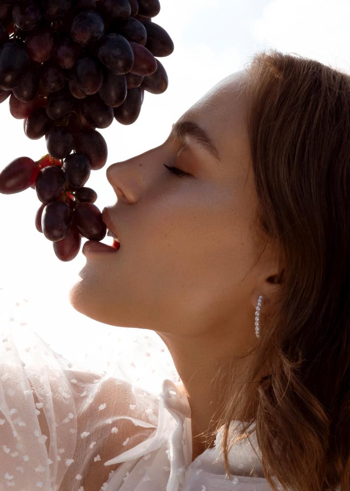 Eine Frau mit geschlossenen Augen, die eine Traube von einem Büschel nascht, während sie ein weißes, durchsichtiges Oberteil trägt.