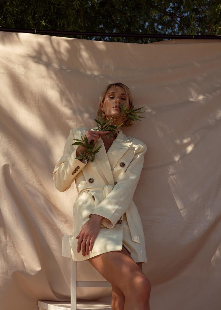 Eine Frau in einem weißen Blazer hält einen kleinen Zweig mit Blättern, während sie auf einem Stuhl sitzt.