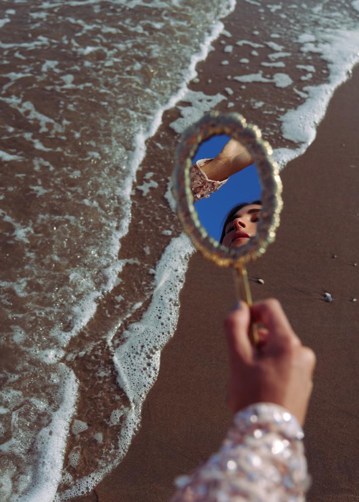 Eine Hand hält einen kleinen Spiegel über den Strand, der das Gesicht einer Frau reflektiert, während Wellen im Hintergrund brechen.