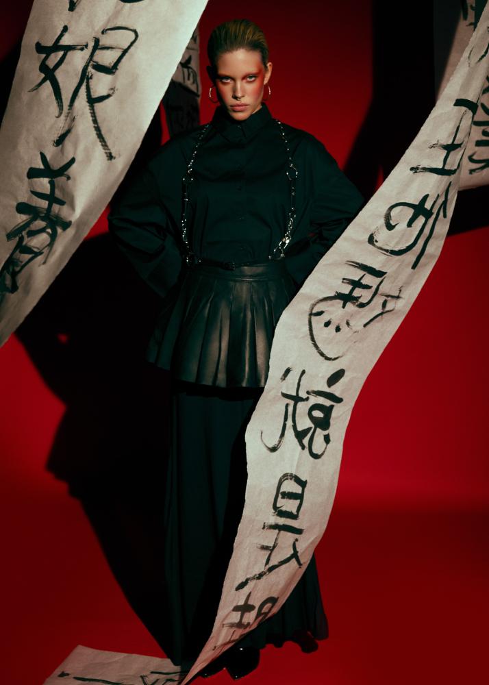 Eine Frau in einem dunklen, strukturierten Outfit mit asiatischen Schriftzeichen auf Zwei Stoff-Bannern.