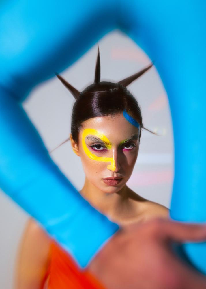 Eine Frau mit dramatischem Make-up und einer futuristischen Frisur, die durch die blauen Arme einer anderen Person blickt.