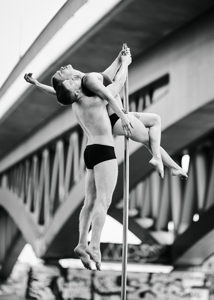 Zwei Pole-TänzerInnen in einer dynamischen Pose unter einer Brücke, die Kraft und Anmut zeigt.